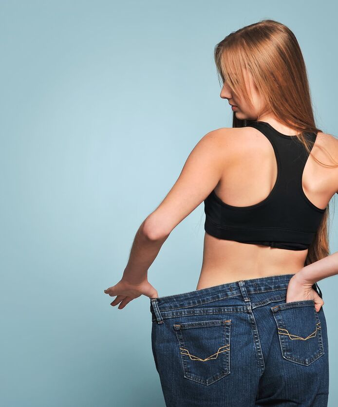 A young woman holds out the waistband of a loose pair of jeans, indicating successful weight loss.